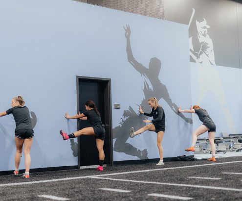A team of athletes are gathered alongside a wall doing leg movement drills.