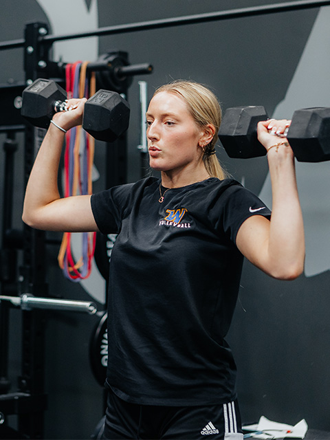 An athlete lifts a pair of dumbbells over her shoulders.