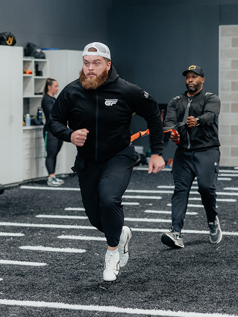 A sports performance coach guides an athlete through a sprint drill on the turf.