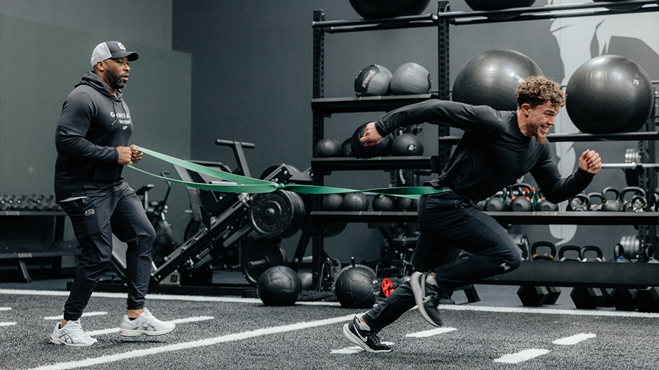 An athlete runs across the turf while a sports performance coach follows, adding resistance to the athlete's sprint.