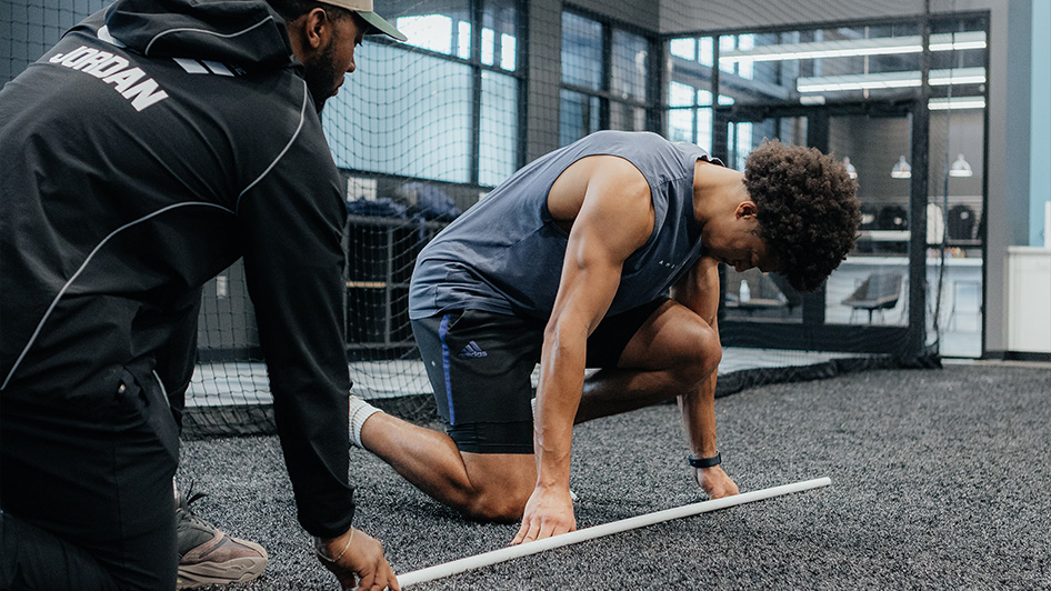 An athlete crouches on the turf while a sports performance coach guides him through a drill.