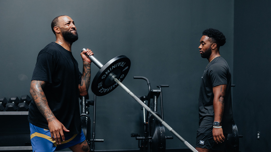 An athlete does an arm strengthening exercise using a weight while a sports performance coach looks on.